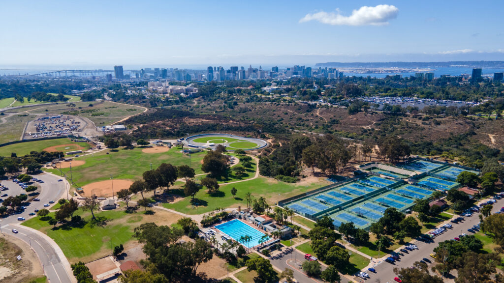 High-altitude aerial drone shot of the Morley Field Sports Complex in Balboa Park, featuring the public swimming pool, tennis courts, baseball diamonds, and the San Diego city skyline in the distance.